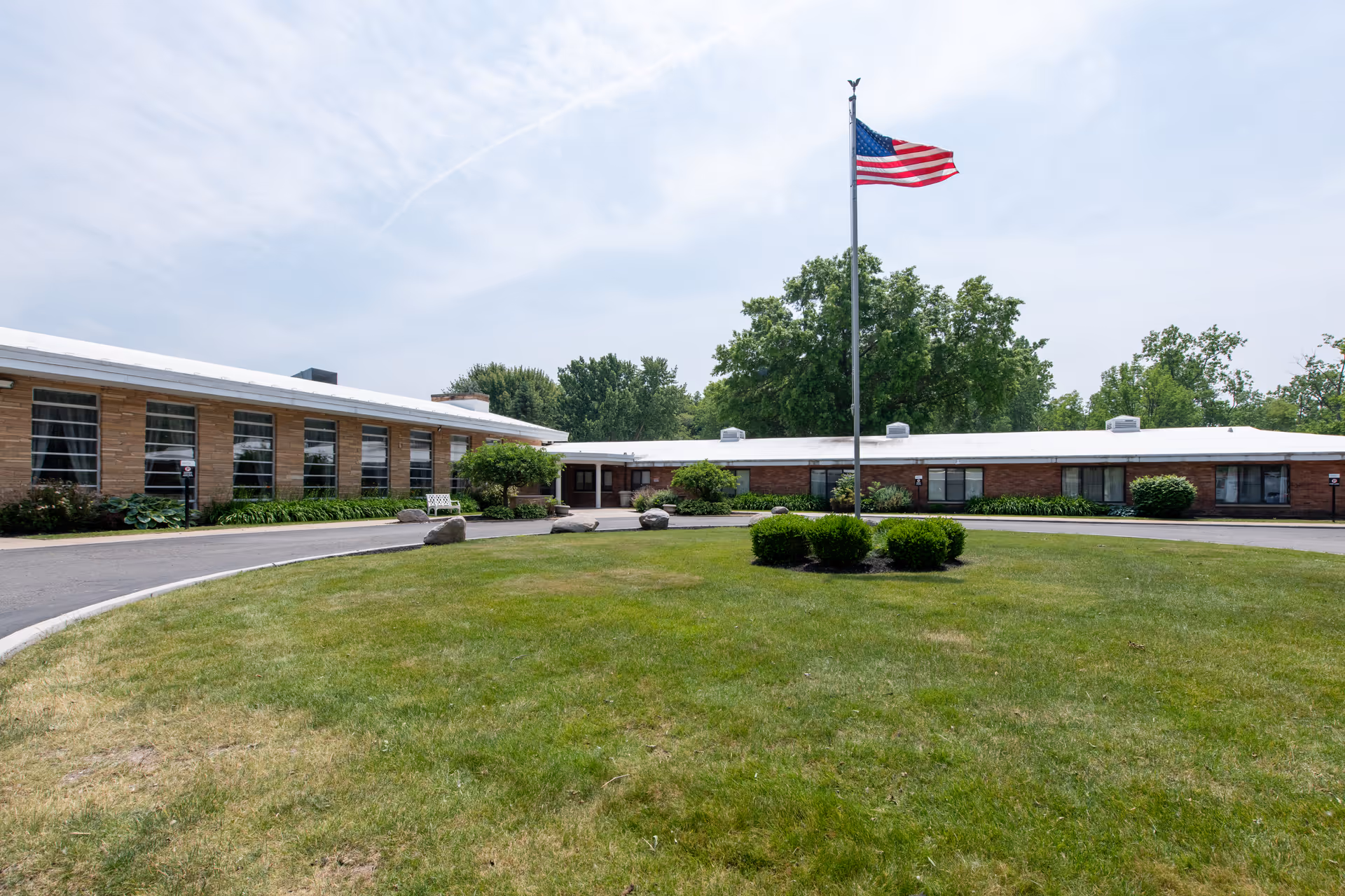 Single-story senior living facility building with a circular driveway, central flagpole flying an American flag, and a grassy courtyard.