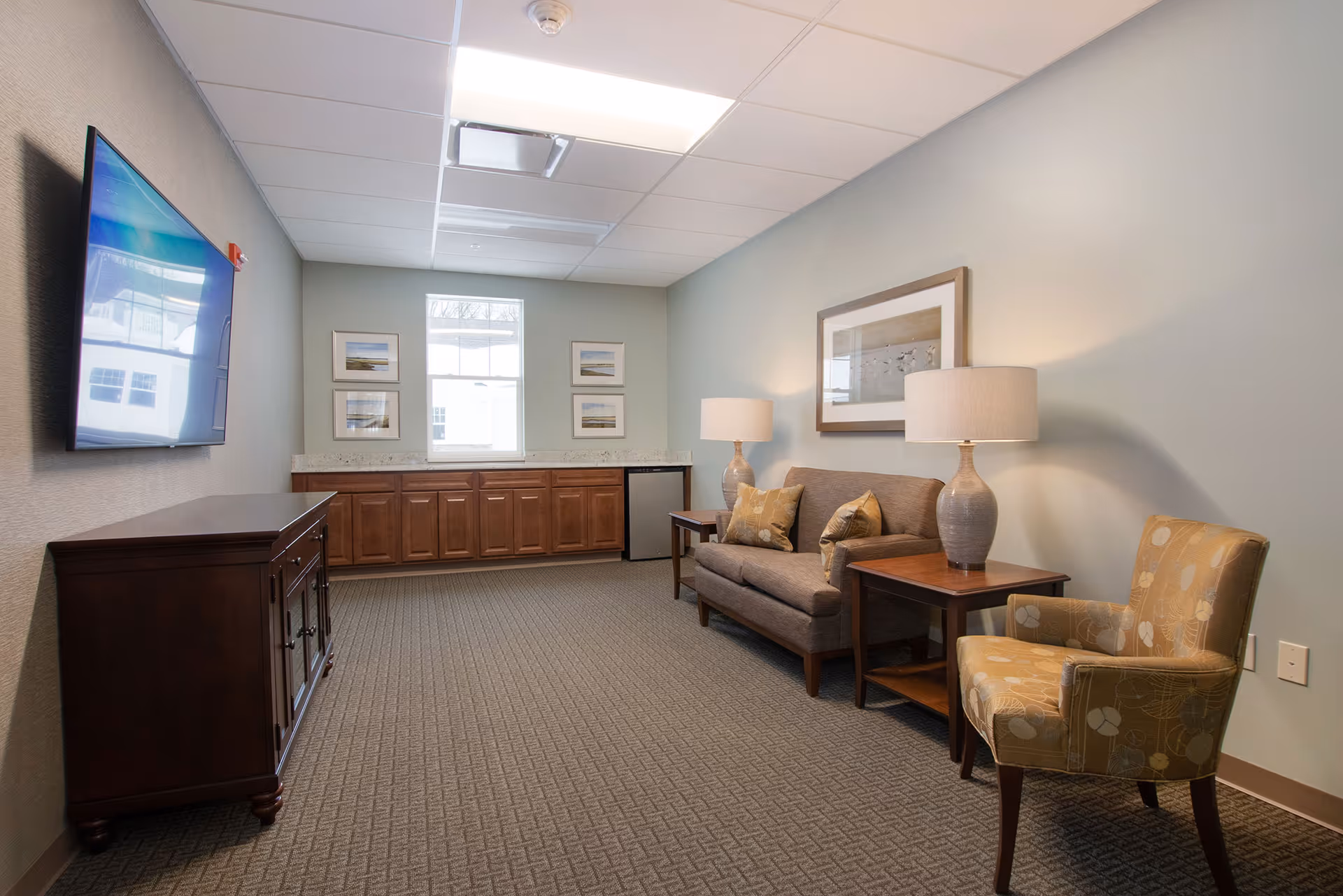 A cozy living room area with a gray sofa, two side tables each with a lamp, a patterned armchair, a wall-mounted flat screen TV, wooden cabinets along the back wall, and framed artwork on the walls.