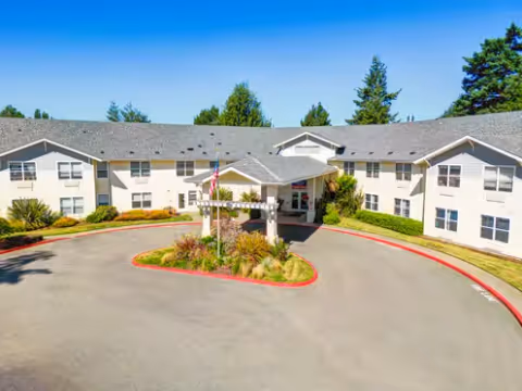 Front exterior view of a two-story senior living facility named Bayside Terrace by Cogir, featuring a circular driveway with landscaped greenery in the center and surrounded by trees under a clear blue sky.