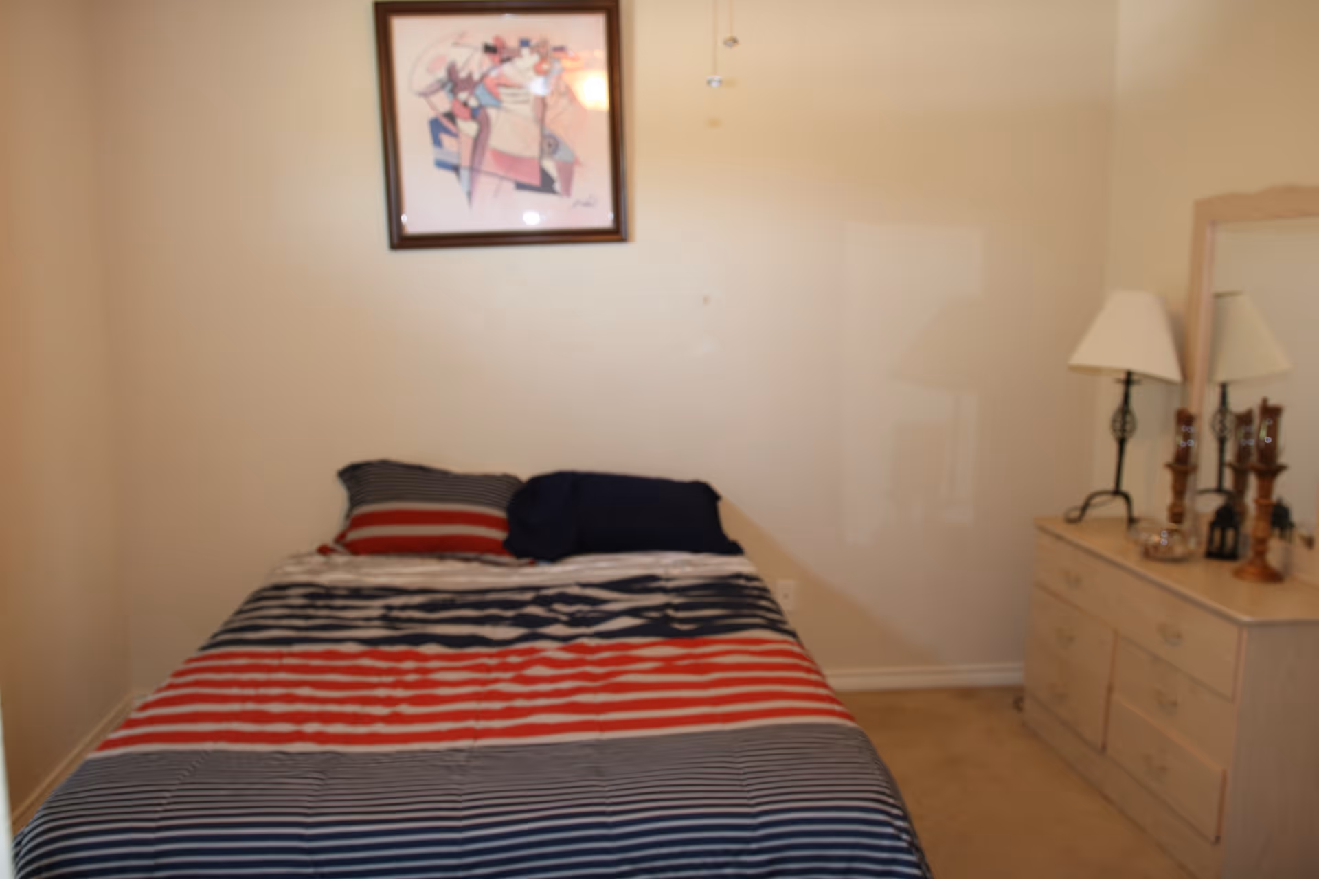 A small bedroom with a bed covered in a striped red, white, and blue comforter and two pillows. There is a framed abstract artwork hanging on the wall above the bed. To the right of the bed is a light-colored dresser with a mirror, a table lamp, and decorative items on top.
