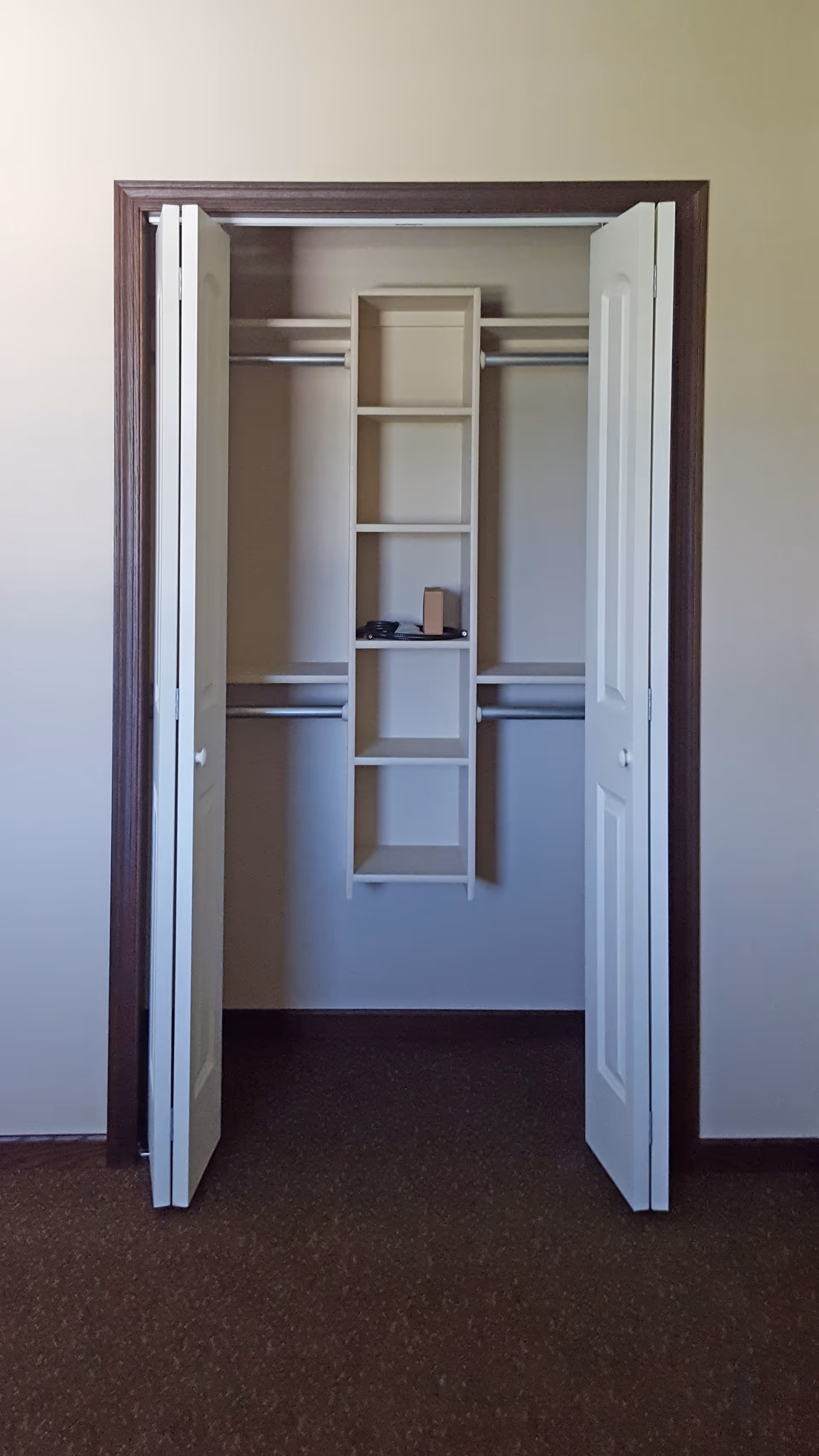An empty closet with white double doors open, revealing a built-in shelving unit in the center and two hanging rods on each side. The closet has beige walls and a brown carpeted floor.