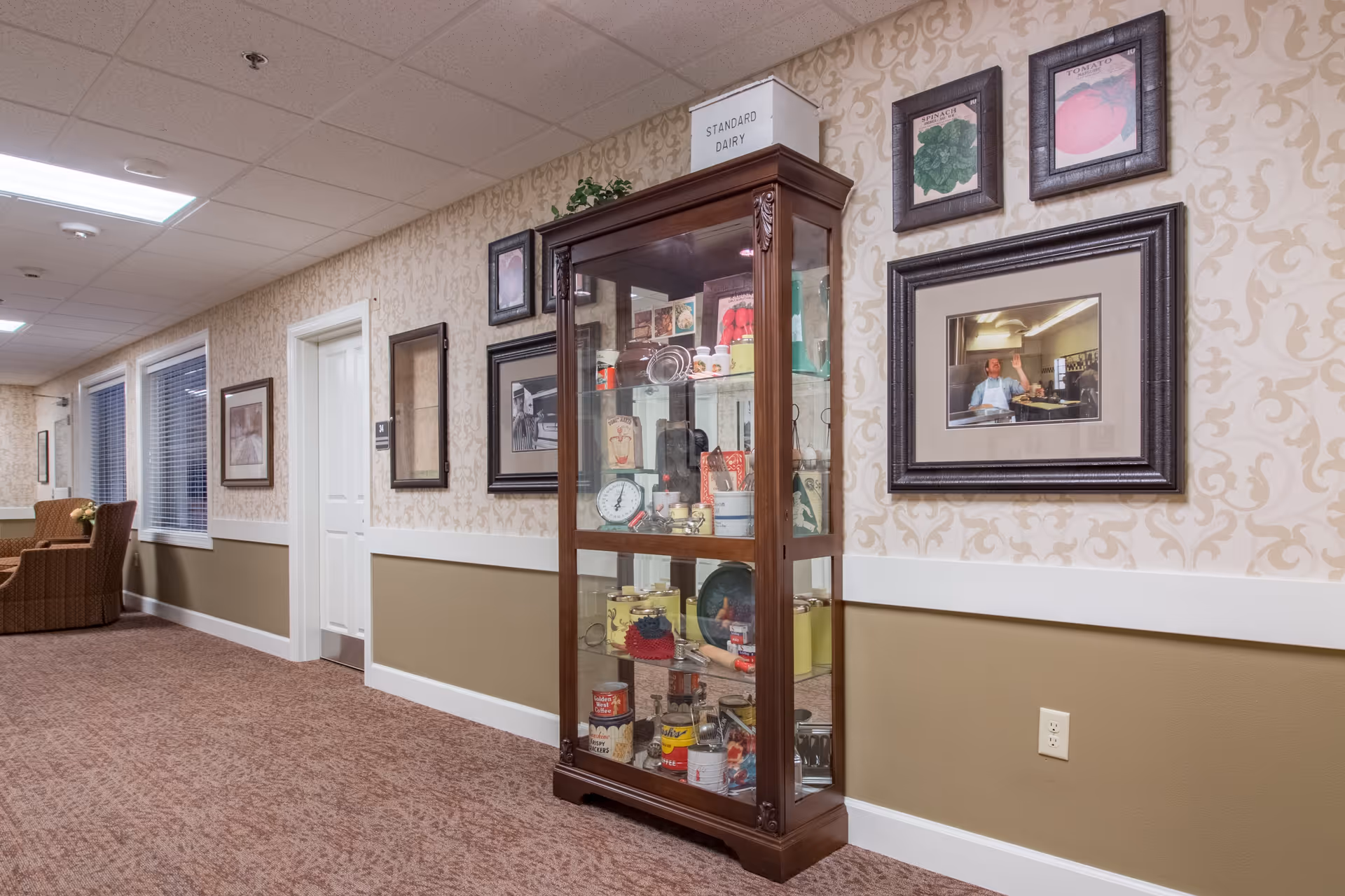 A hallway in a senior living facility with patterned wallpaper and carpeted floor. There is a wooden display cabinet filled with vintage items such as cans, scales, and kitchenware. Several framed pictures and artwork hang on the walls. At the end of the hallway, there are windows with blinds and a seating area with armchairs.