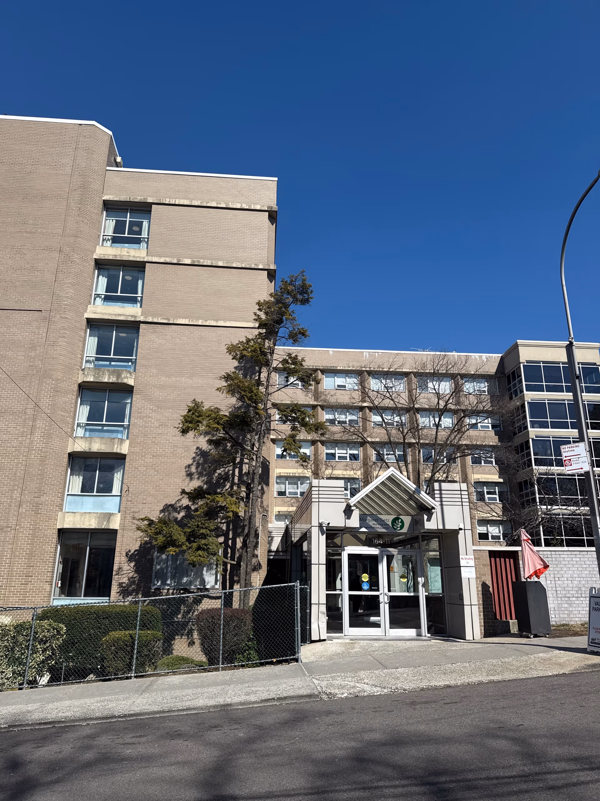 Exterior view of the Margaret Tietz Nursing and Rehabilitation Center building under a clear blue sky. The multi-story building has beige brick walls and several windows. There is a main entrance with a small awning and glass doors, surrounded by some trees and bushes. A street lamp and a parking sign are visible near the entrance.