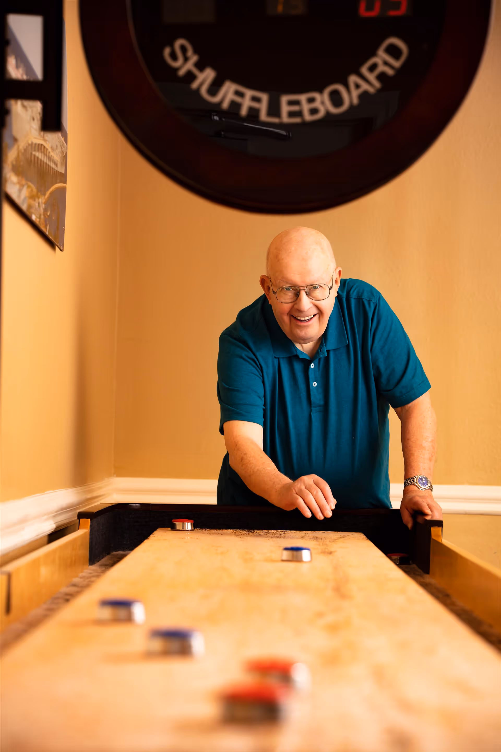 An elderly man wearing glasses and a teal polo shirt is smiling and playing shuffleboard indoors. The shuffleboard table is in the foreground with several pucks on it. A shuffleboard scoreboard is visible on the wall behind him.