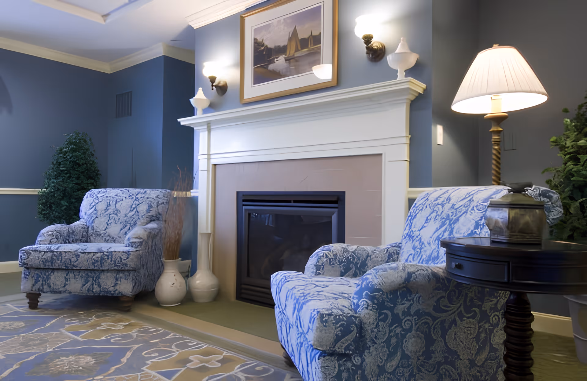 A cozy living room area with two blue and white patterned armchairs facing a white fireplace. Above the fireplace is a framed painting of sailboats on water. The walls are painted blue with white trim, and there are two wall sconces providing light. A floor lamp with a white shade stands next to one armchair, which is beside a dark wooden side table holding a decorative item. There are also some potted plants and vases near the fireplace.
