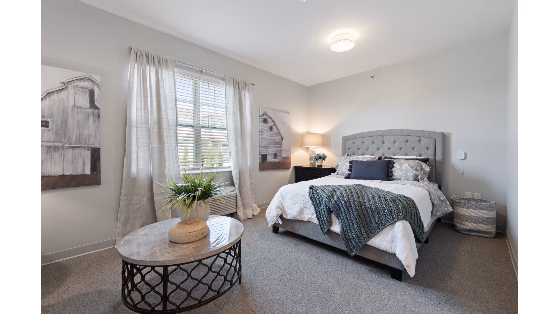 A neatly arranged bedroom with a gray upholstered bed featuring white and patterned bedding, a dark blue throw blanket, and decorative pillows. There is a round marble-top coffee table with a potted plant in the foreground. The room has light gray walls, a window with white blinds and light curtains, two paintings of barns on the walls, a black nightstand with a lamp and a small plant, and a striped basket on the floor.