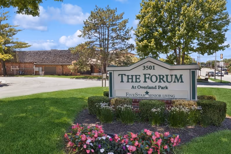 Outdoor view of the entrance sign for The Forum at Overland Park senior living facility, surrounded by green grass, bushes, and colorful flowers, with a building and trees in the background under a partly cloudy sky.