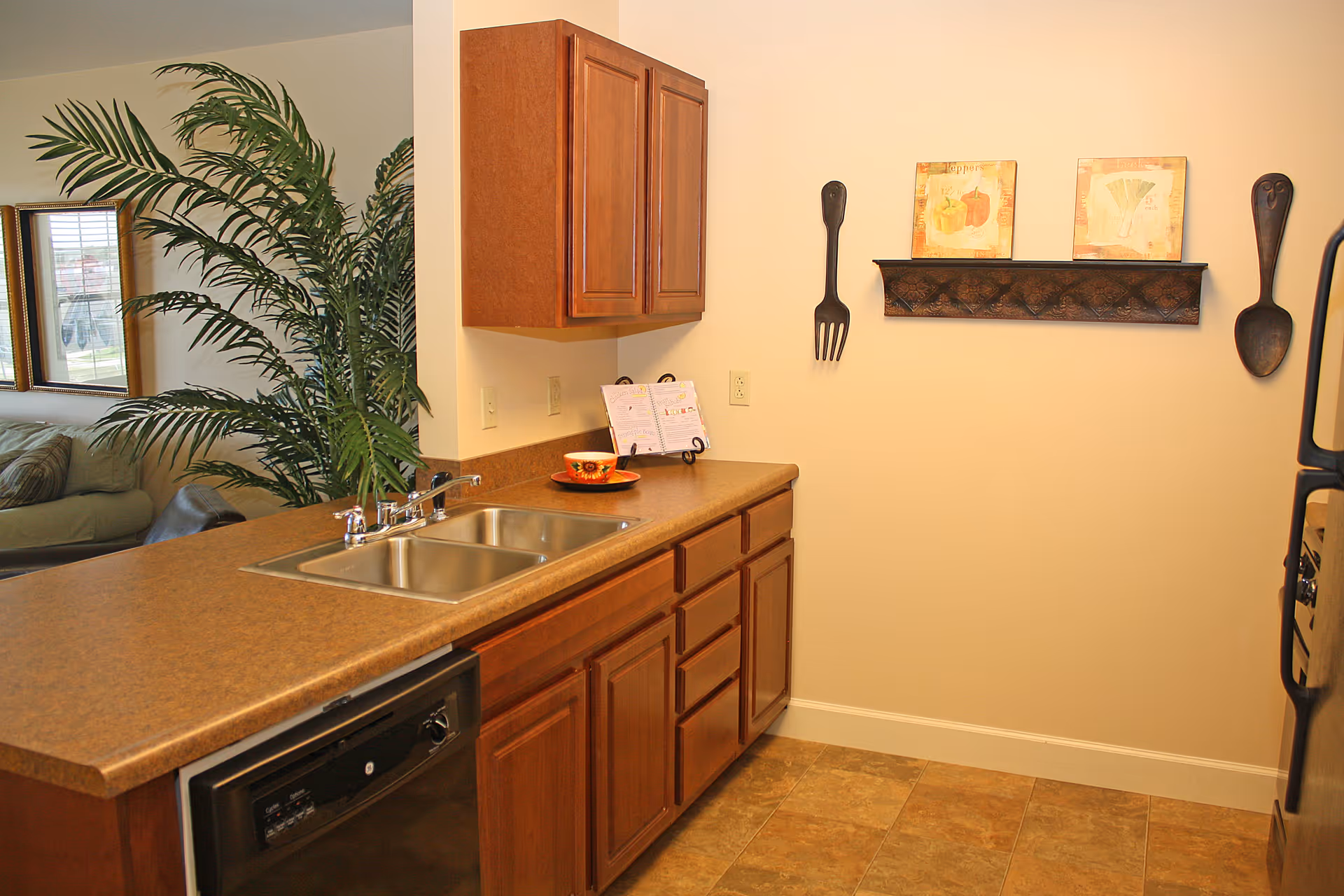 A kitchen area with a brown countertop featuring a double sink and a dishwasher underneath. There are wooden cabinets above and below the counter. On the wall, there is a decorative shelf with two framed pictures and large fork and spoon wall hangings. A large green plant and part of a living room with a couch and window are visible in the background.