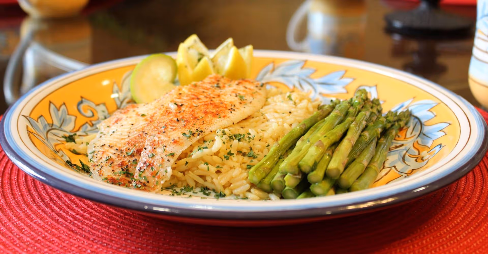 A plate of food featuring a seasoned grilled fish fillet, a serving of rice, steamed asparagus, and lemon wedges, placed on a decorative yellow and blue plate on a red placemat.