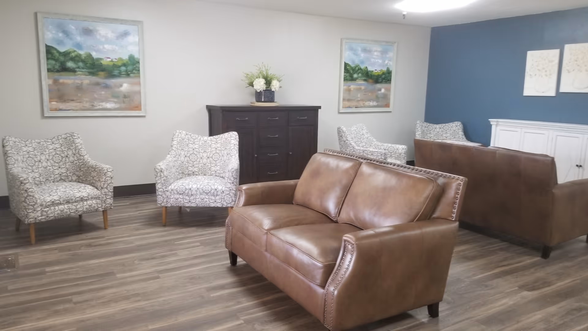 A cozy living room area with two brown leather sofas and four patterned armchairs arranged on a wood floor. The walls are decorated with landscape paintings and floral artwork. A dark wooden cabinet with a flower arrangement sits against one wall.