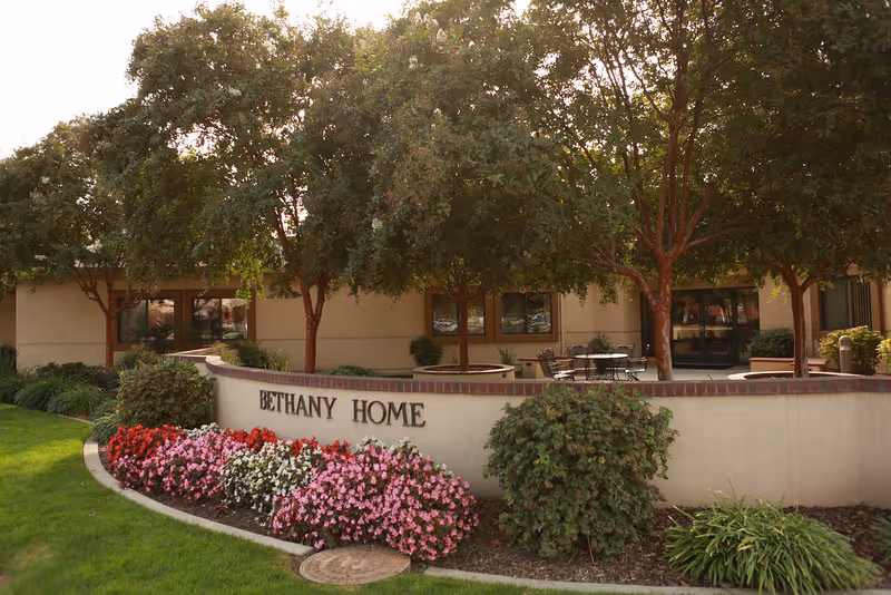 Exterior view of Bethany Home facility showing a curved low wall with the name 'Bethany Home' on it, surrounded by colorful flower beds and green shrubs. Behind the wall are several trees and a building with windows and a patio area with tables and chairs.