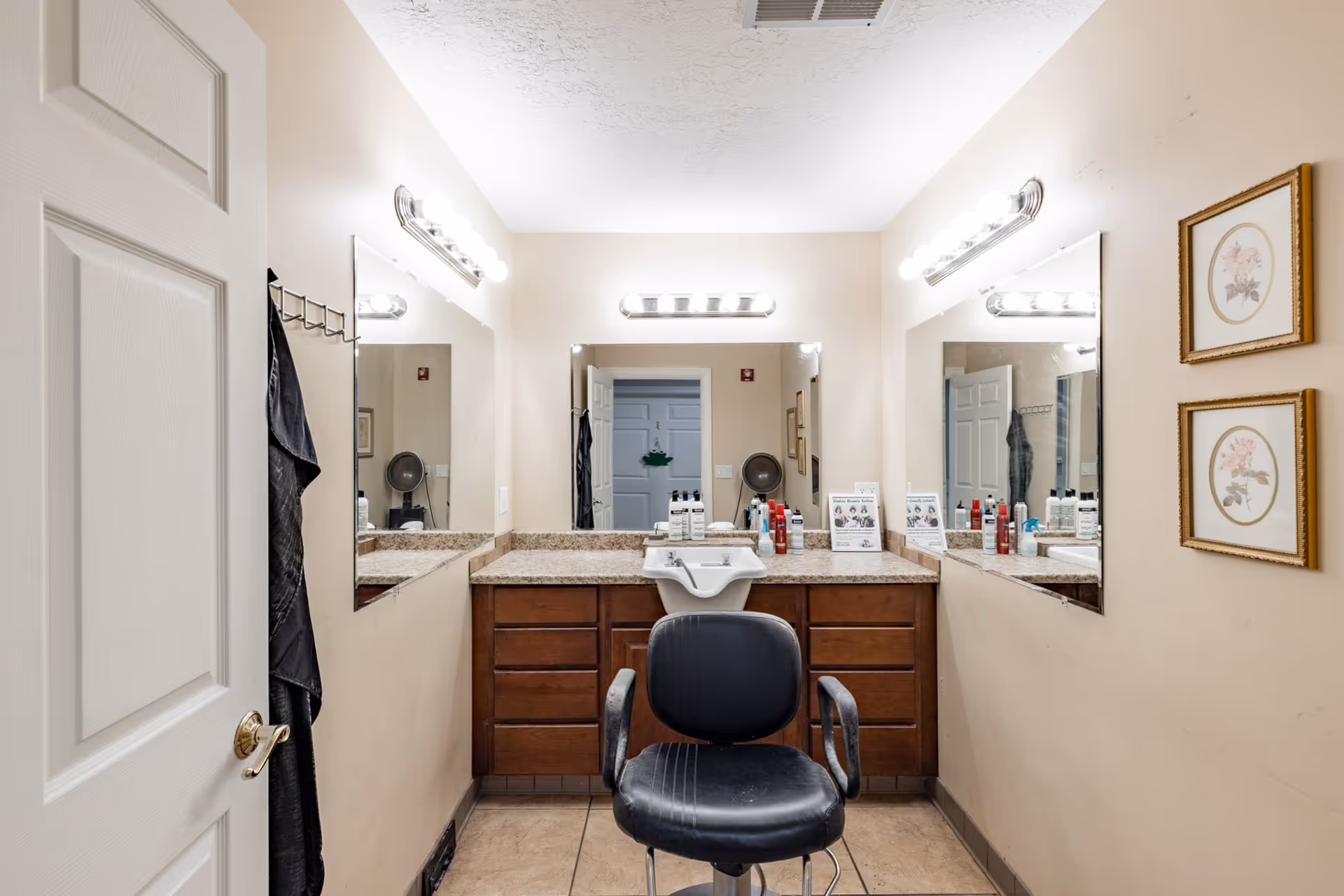 Interior view of a beauty salon area in a senior living facility with a black salon chair in front of a counter with a sink, various hair care products, and three large mirrors on the walls. The room has beige walls, tile flooring, and bright vanity lights above the mirrors. Two framed floral pictures hang on the right wall.