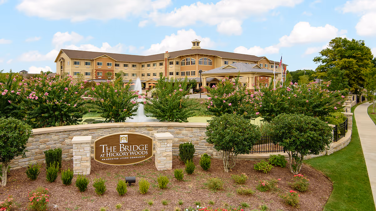 Exterior view of The Bridge at Hickory Woods assisted living community building with a landscaped garden, flowering bushes, a water fountain, and a stone sign displaying the facility's name.