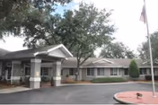 Front entrance of a single-story senior living facility with a covered porte-cochère, trees, and a flagpole.