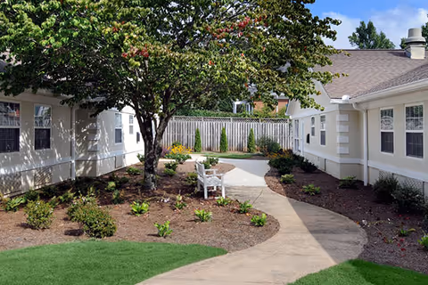 Courtyard with a winding concrete path between two single-story buildings, landscaped planting beds, a tree and a white bench.