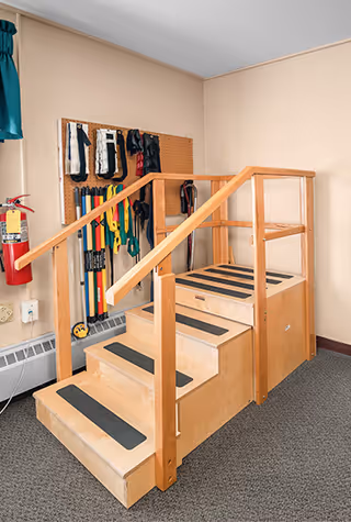 A wooden rehabilitation staircase with handrails on both sides and non-slip strips on each step, situated indoors against a beige wall. Behind the staircase, various exercise and therapy equipment hang on a pegboard. A fire extinguisher is mounted on the wall to the left.