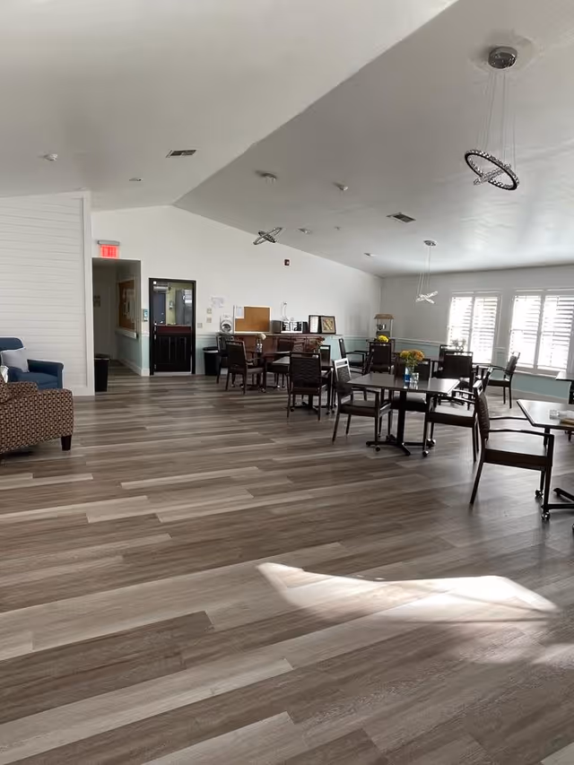 Spacious communal dining/activity room with tables and chairs, large shuttered windows, pendant lighting, and wood-look flooring.