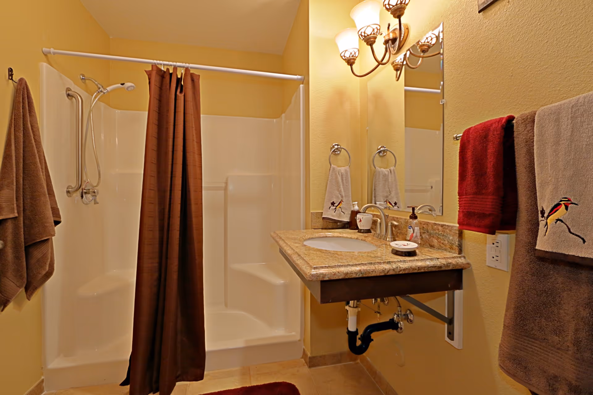 A bathroom with a walk-in shower featuring a brown shower curtain and grab bars. There is a wall-mounted sink with a granite countertop, a mirror above it, and several towels hanging on towel racks. The walls are painted yellow and the lighting fixture above the mirror has three lights.