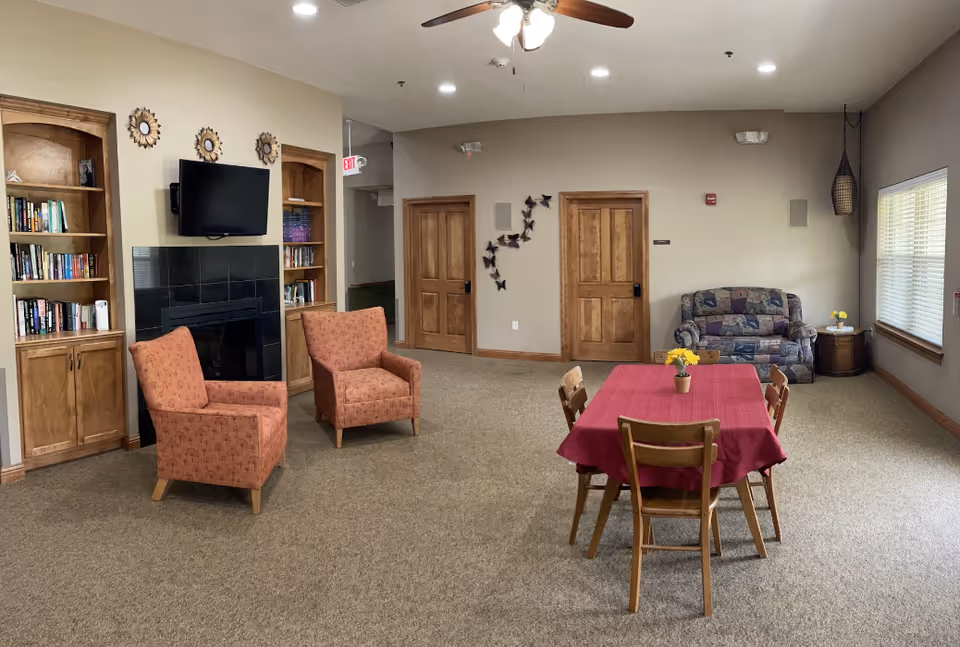 Sunrise Meadows common lounge with two pink armchairs, fireplace, shelves, and a red tablecloth.