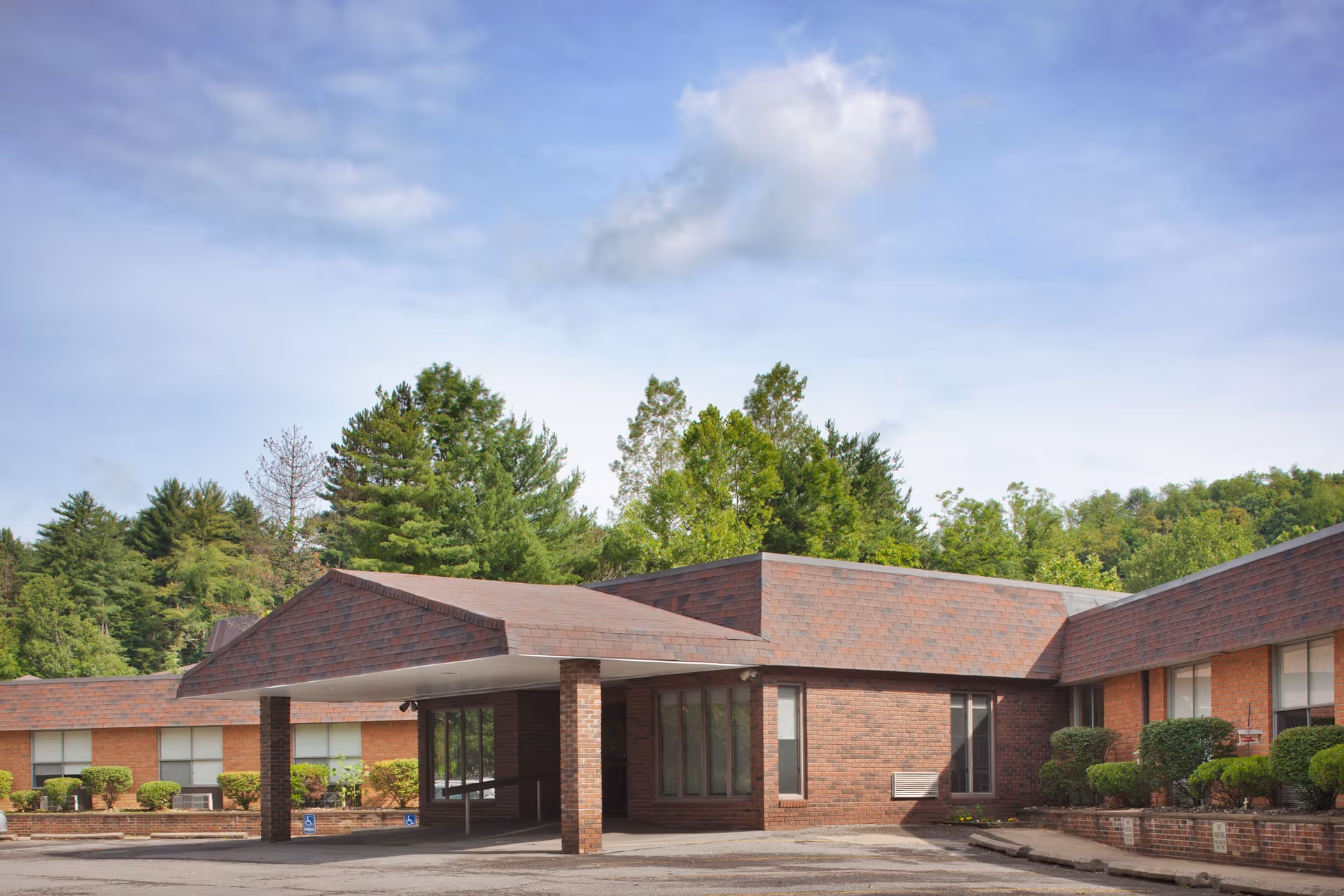 Exterior view of a single-story brick building with a covered entrance supported by brick columns, surrounded by greenery and trees under a partly cloudy sky.