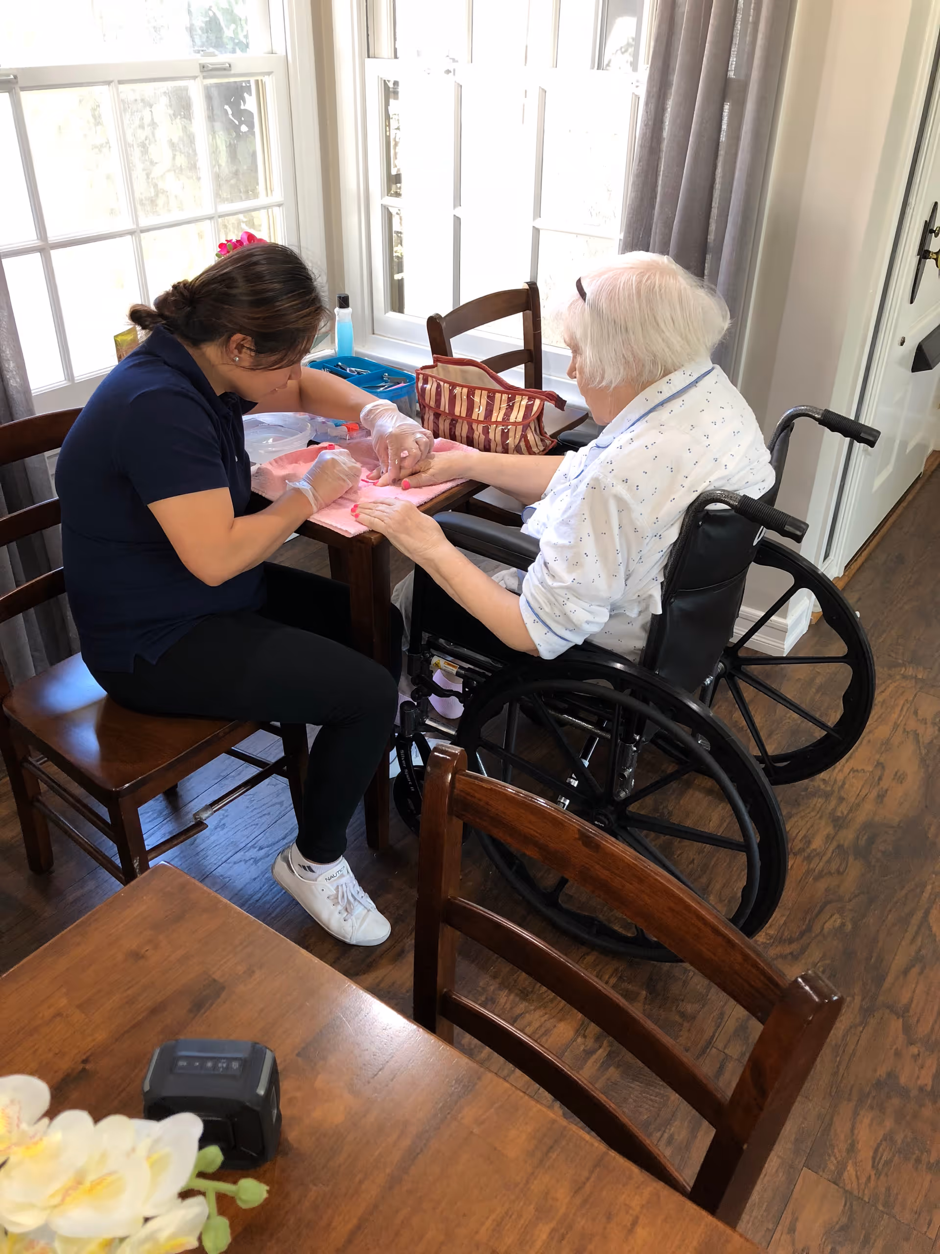 A caregiver is providing a manicure to an elderly woman in a wheelchair at a wooden table near a window with white curtains. The caregiver is wearing gloves and a navy blue shirt, while the elderly woman is dressed in a white shirt with small patterns. The room has wooden flooring and several wooden chairs.