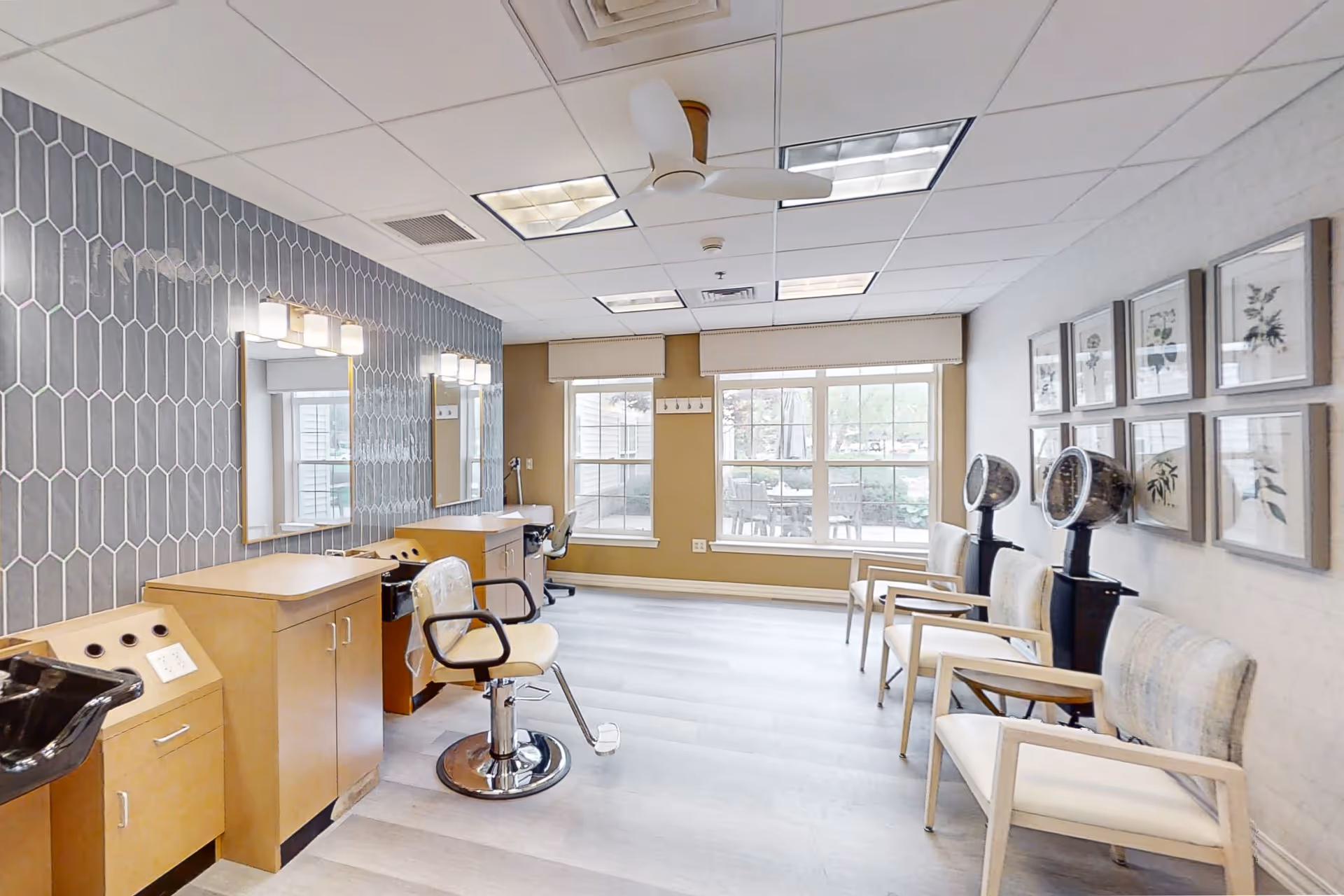 Interior of a senior living facility hair salon with styling chairs, mirrors, hair washing station, and hair dryers. The room has large windows letting in natural light, framed botanical artwork on the wall, and a ceiling fan.