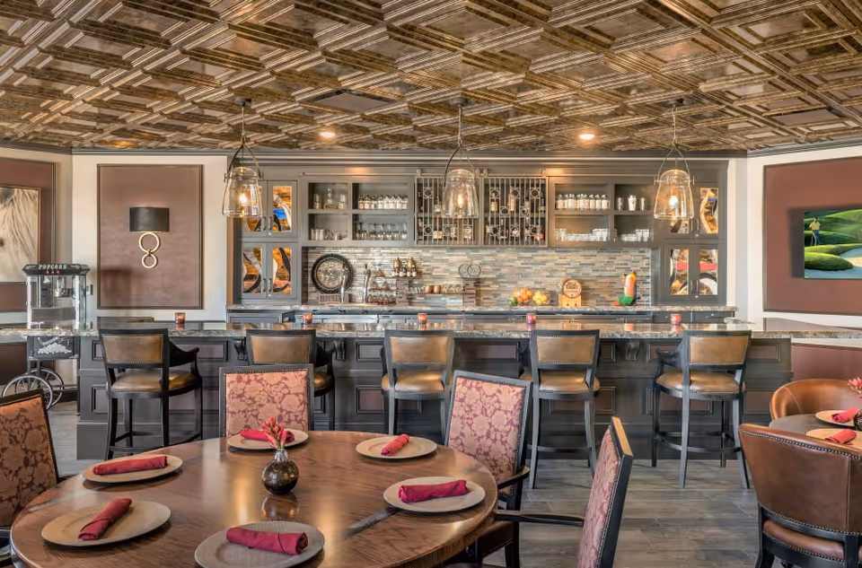 A stylish dining area with a round wooden table set with plates and red napkins, surrounded by upholstered chairs. In the background, there is a bar counter with high chairs, shelves stocked with glasses and bottles, and three hanging pendant lights. The ceiling has a decorative wooden pattern, and the walls feature framed artwork and a popcorn machine on the left side.