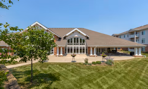 Exterior view of a single-story building with large windows and a sloped roof, surrounded by a well-maintained lawn and a few small trees under a clear blue sky.