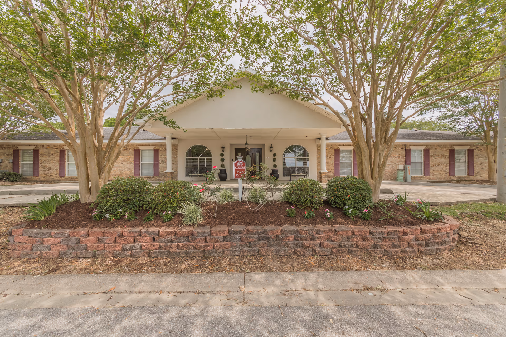 Front exterior view of Rosewood Assisted Living facility showing a single-story brick building with a covered entrance supported by white columns. There are two large trees and landscaped bushes and flowers in front of the building. A sign near the entrance reads 'Rosewood Main Office'.