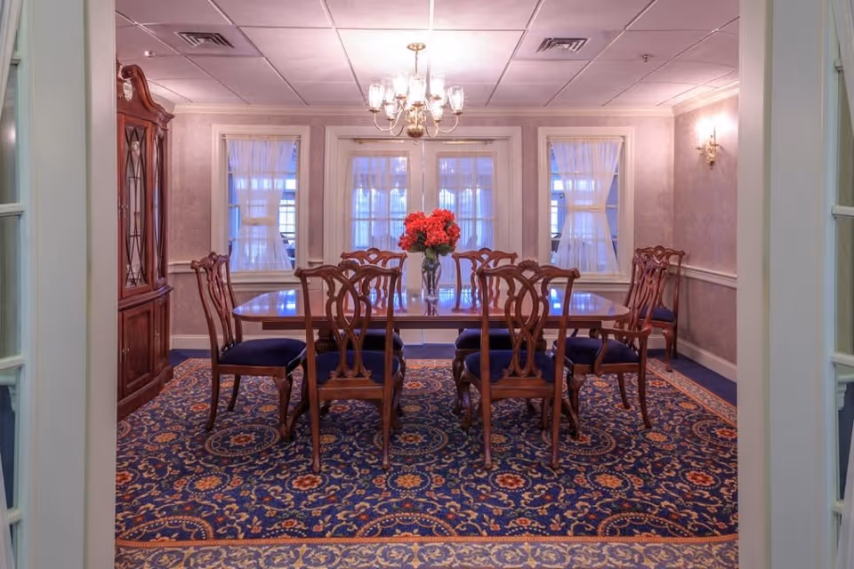 A formal dining room with a wooden dining table surrounded by eight matching wooden chairs with blue cushions. A vase with red flowers is placed in the center of the table. The room features a patterned blue carpet, light purple walls with white trim, three windows with sheer white curtains, a chandelier hanging from the ceiling, and a wooden china cabinet on the left side.