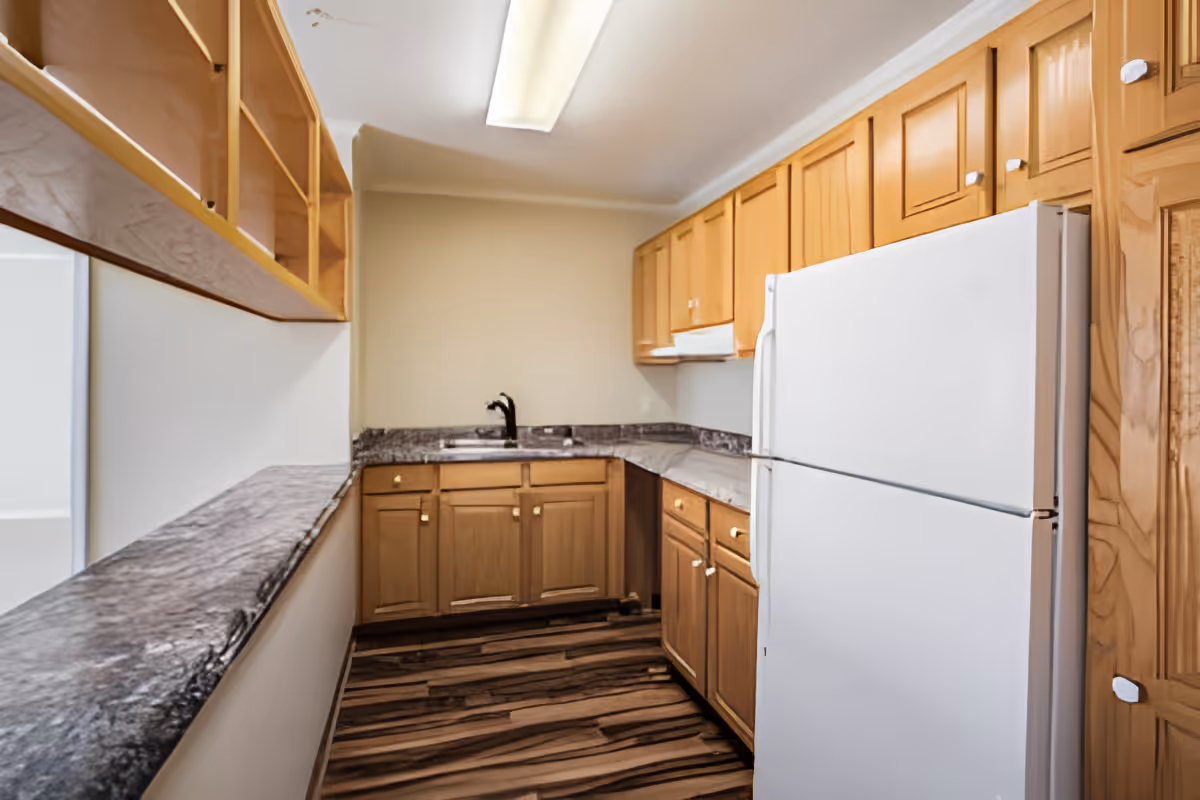 A small kitchen with wooden cabinets, a white refrigerator, a sink with a black faucet, and marble-patterned countertops. The floor has a wood-like pattern, and there is a fluorescent ceiling light.