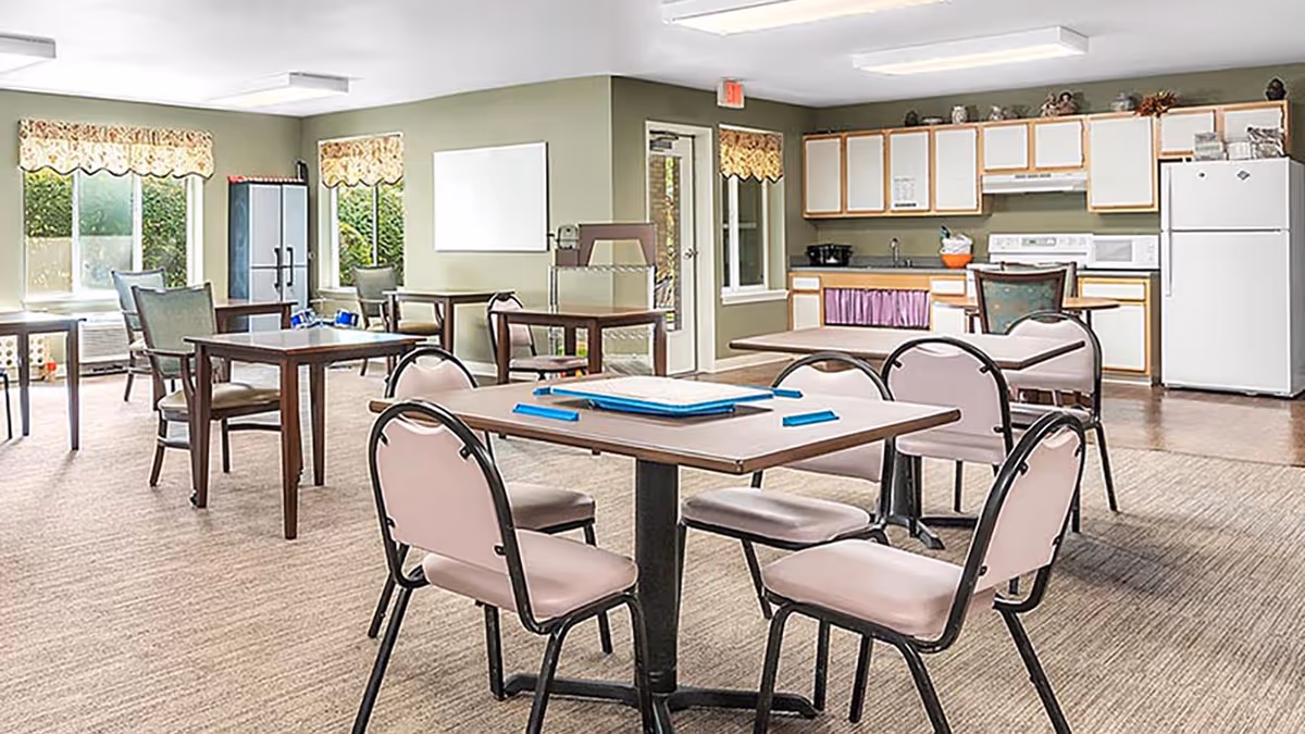 Bright communal dining/activity room with several tables and chairs and a kitchenette along the back wall.