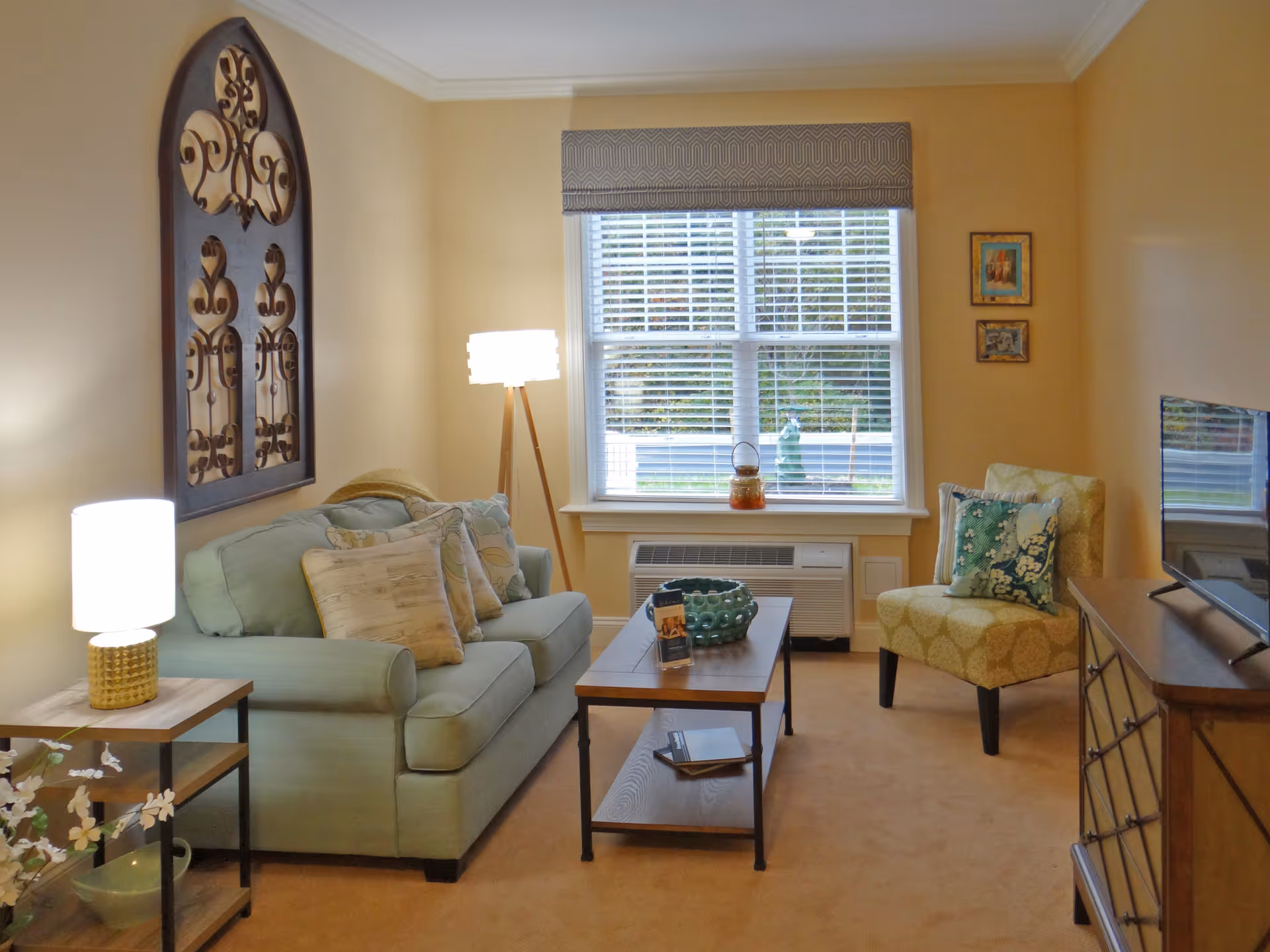 A cozy living room with a light green sofa adorned with multiple pillows, a wooden coffee table with decorative items, a patterned armchair with a cushion, a wooden side table with a lamp, a standing lamp, and a window with blinds and a valance letting in natural light. The walls are painted a warm beige color and decorated with framed artwork and a large decorative wall piece.