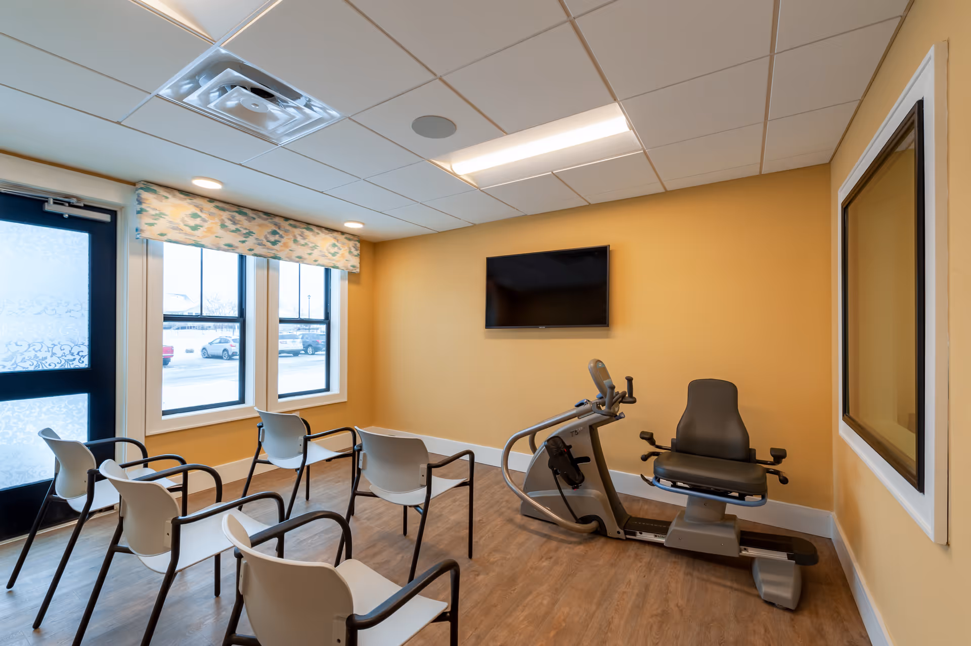 A small exercise room with a recumbent stationary bike, six white chairs with black armrests arranged facing a wall-mounted flat screen TV. The room has yellow walls, wood flooring, a window with a patterned valance, and a door with frosted glass panels.