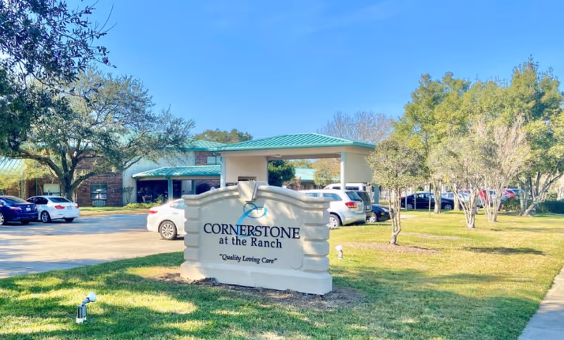 Exterior view of Cornerstone at the Ranch Bethany Rehabilitation facility with a large sign in the foreground displaying the facility name and slogan 'Quality Loving Care'. Several cars are parked near the building, which has a green roof and is surrounded by trees and grass under a clear blue sky.