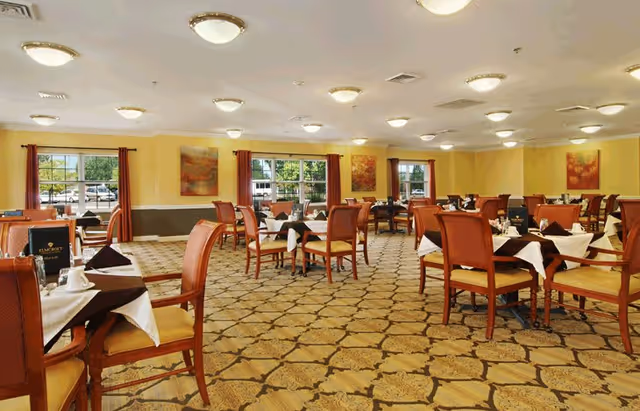 A spacious dining room with multiple tables covered with white tablecloths and brown napkins, surrounded by wooden chairs with yellow cushions. The room has large windows with red curtains, yellow walls with paintings, and a patterned carpeted floor. Ceiling lights illuminate the area.