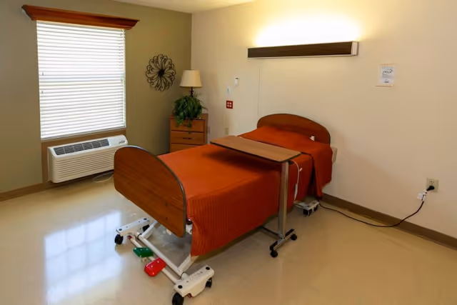 A simple, clean room with a single hospital-style bed covered with an orange blanket. The bed has wheels and an adjustable overbed table. There is a small wooden dresser with a lamp and a plant on top, a window with blinds, and a decorative wall piece. The floor is tiled and the walls are painted in neutral tones.