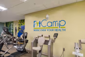 An indoor fitness room with exercise machines in front of a light green wall bearing a 'FitCamp Rediscover Your Health' sign.