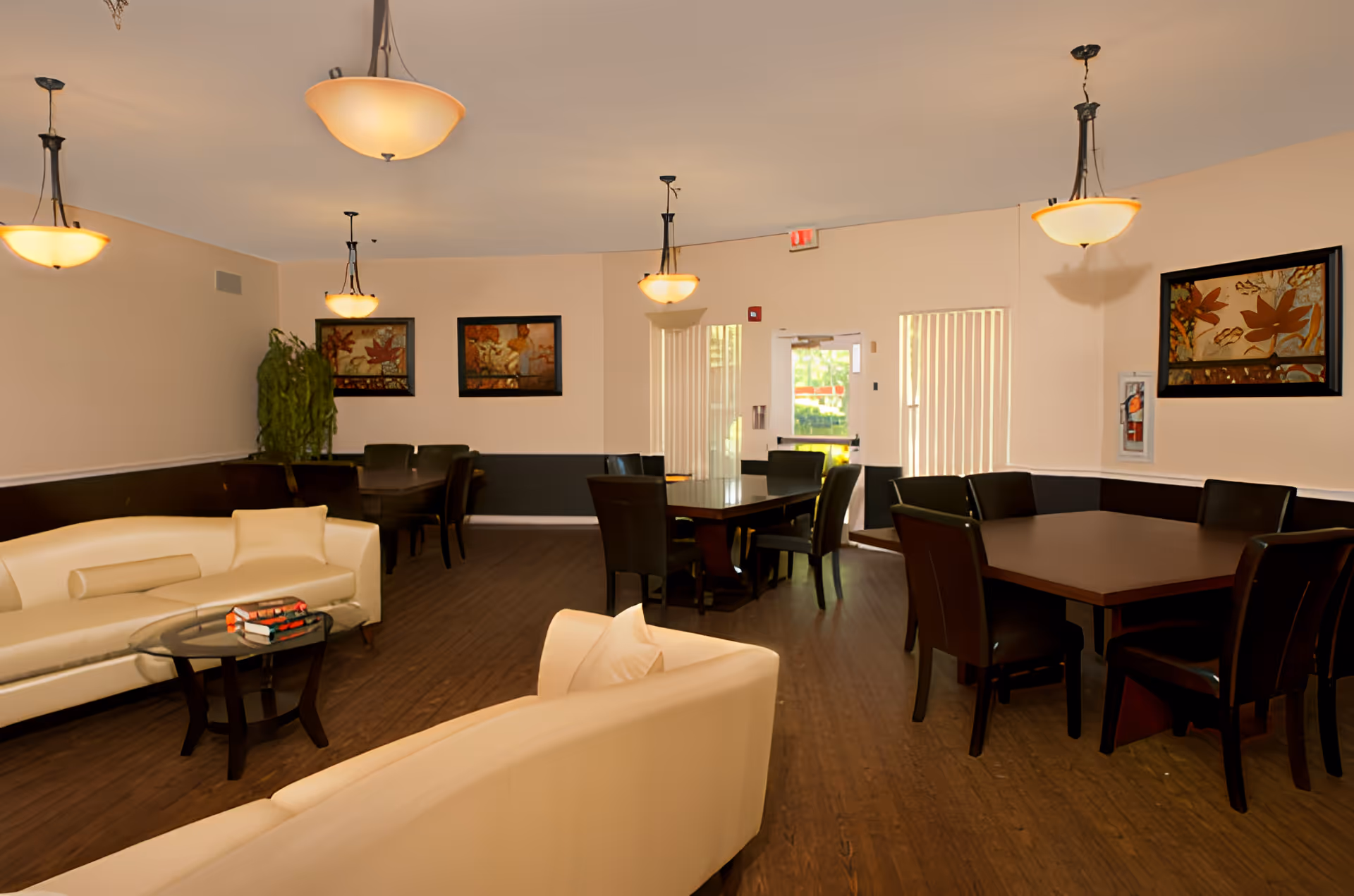 A spacious, well-lit common area in Riverdale Assisted Living featuring cream-colored sofas with pillows, a small glass coffee table with books, several dark wooden dining tables with black chairs, framed artwork on beige walls, hanging pendant lights, and a door with vertical blinds letting in natural light.
