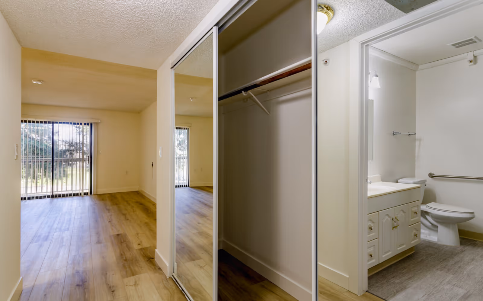 Interior view of a senior living facility room showing a wooden floor, a large sliding glass door with vertical blinds leading outside, a closet with mirrored sliding doors, and a bathroom with a toilet, sink, and grab bars.