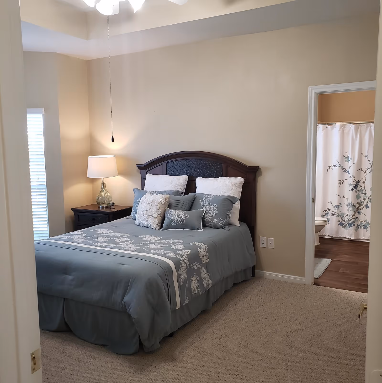 A cozy bedroom featuring a queen-sized bed with a light blue comforter and decorative pillows, a bedside lamp, and a window with blinds. A bathroom is visible through an open door.