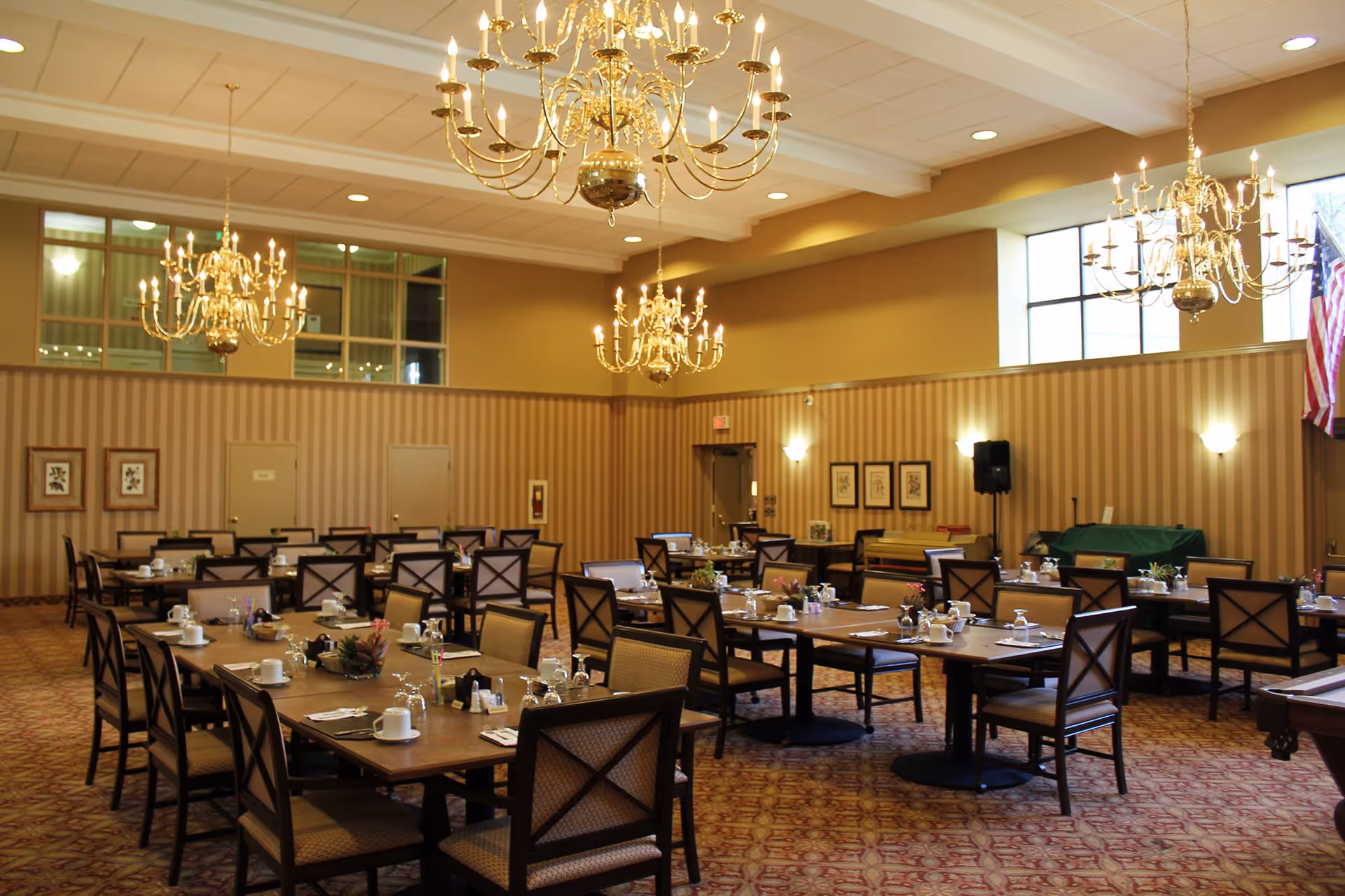 A spacious dining room with multiple rectangular tables set with cups, glasses, and silverware. The room features patterned carpet, striped wallpaper, several framed pictures on the walls, large windows, and multiple ornate chandeliers hanging from the ceiling. An American flag is visible in the corner.