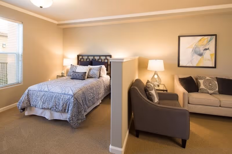 A cozy bedroom with a bed covered in a blue patterned quilt and multiple pillows. Next to the bed is a small nightstand with a lamp. A half wall separates the bedroom from a sitting area that includes a gray armchair and a beige sofa with decorative pillows. A framed abstract painting hangs on the wall above the sofa. The room has beige walls and carpet, and a window with blinds lets in natural light.