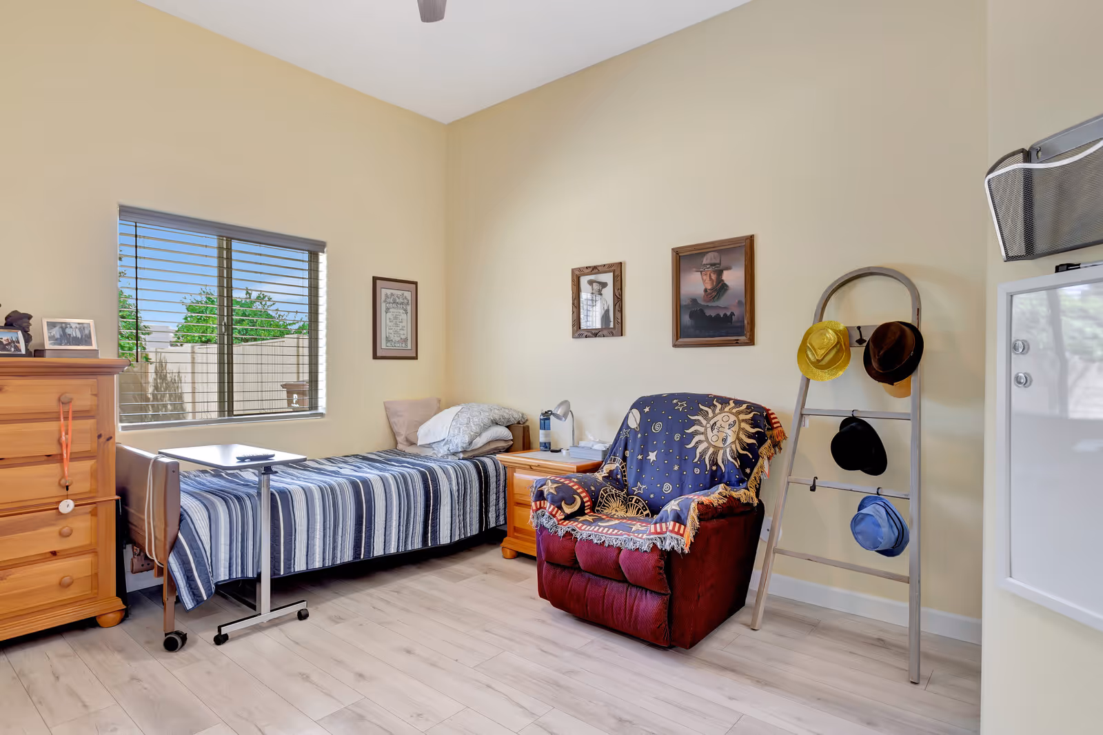 A cozy bedroom in an assisted senior living facility featuring a single bed with a striped blanket, a wooden dresser, a bedside table with a lamp, a comfortable armchair draped with a celestial-themed blanket, and a ladder-style hat rack holding several hats. The room has light-colored walls, a window with blinds showing greenery outside, and framed pictures on the wall.