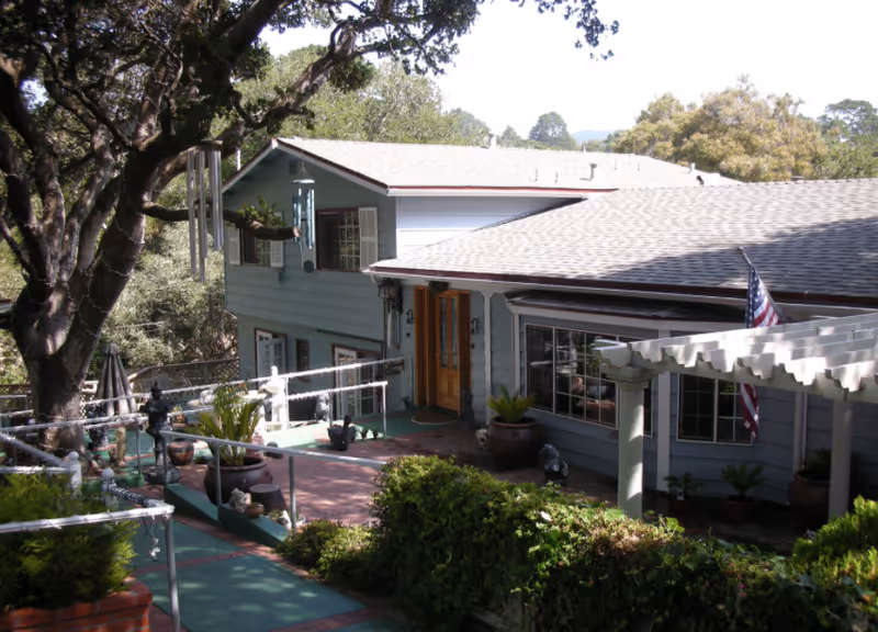 Exterior view of a residential care home building with a ramp and stairs leading to a wooden front door. The building is surrounded by trees and greenery, with an American flag hanging near a white pergola structure.