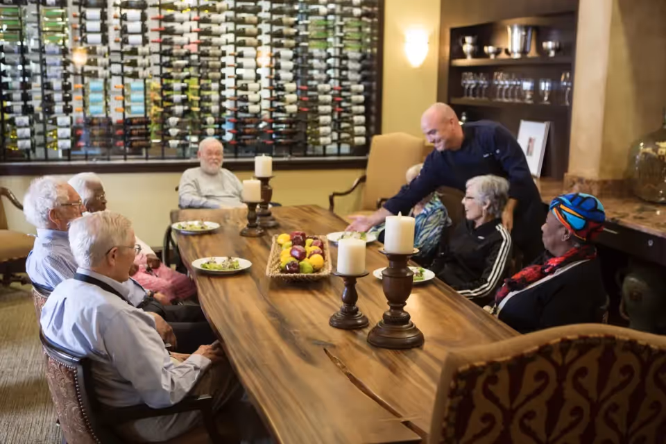 A group of elderly people sitting around a large wooden dining table with plates of food. A staff member is serving or interacting with them. The background features a wall-mounted wine rack and shelves with glassware.