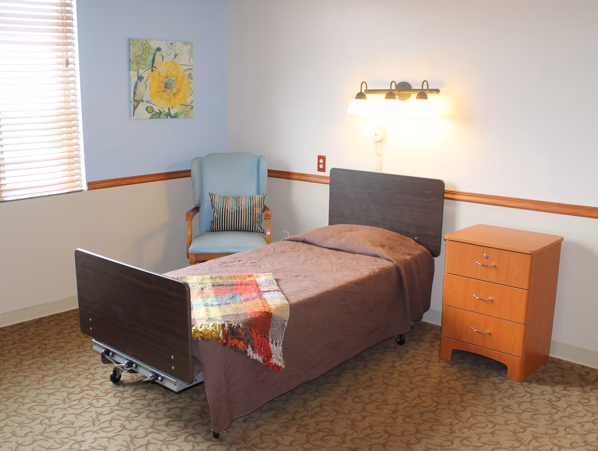 A simple bedroom in a senior living facility with a single bed covered in a brown blanket and a colorful throw at the foot. Next to the bed is a wooden nightstand with three drawers. A light blue armchair with a striped pillow is placed in the corner near a window with blinds. A wall-mounted light fixture is above the bed, and a painting of birds and a yellow flower hangs on the wall.