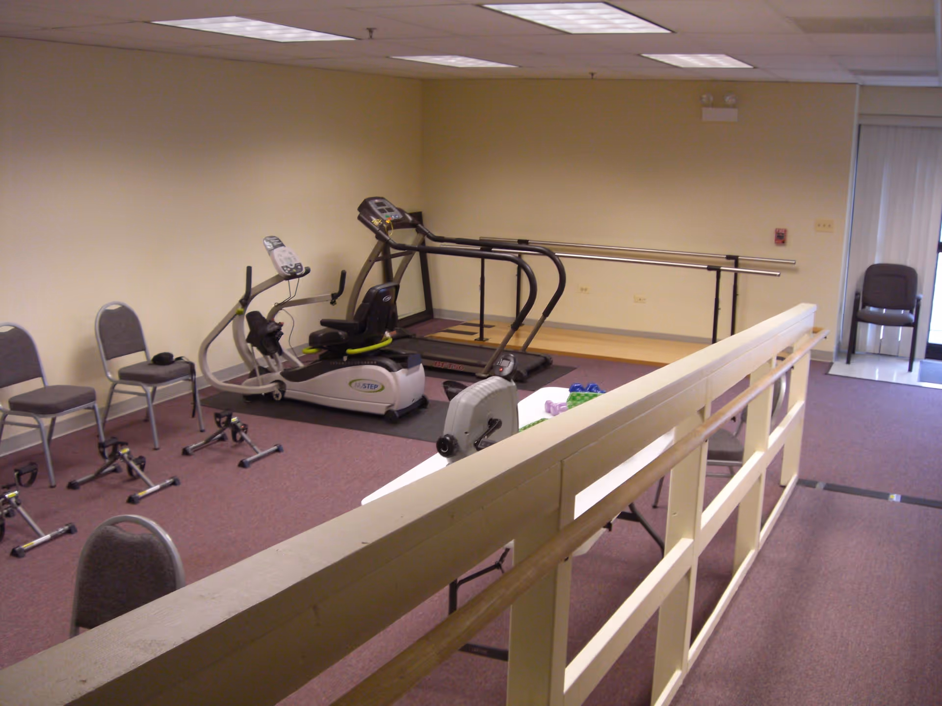An interior exercise/therapy room with a recumbent bike, treadmill, parallel bars, chairs and small pedal exercisers.