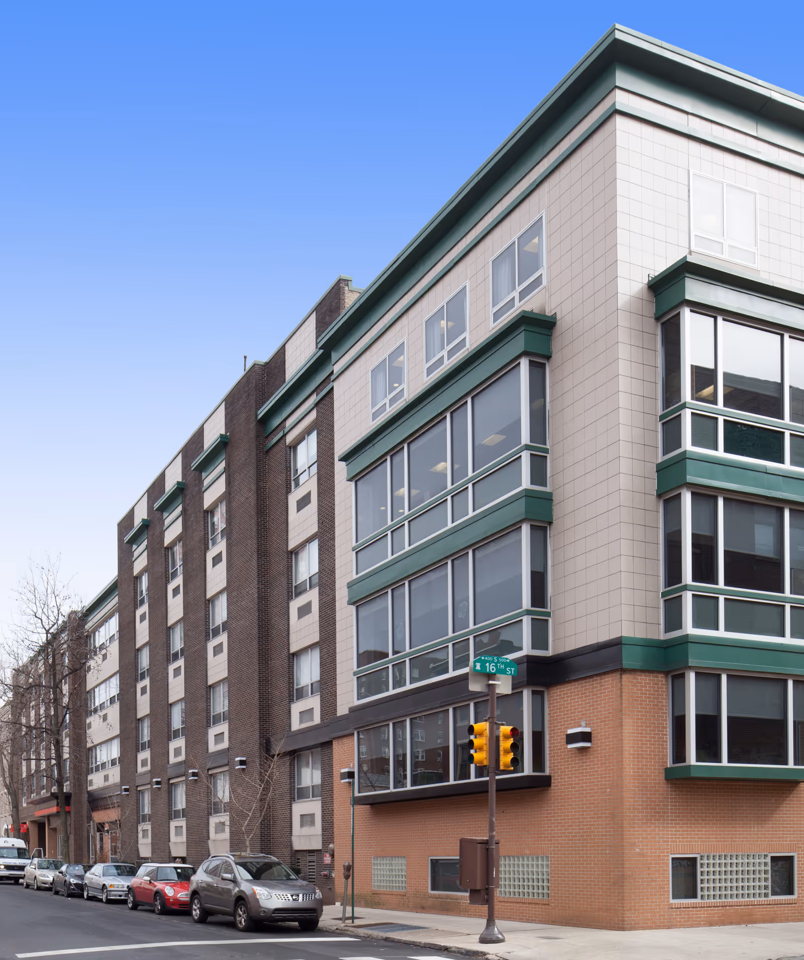 Exterior view of a multi-story building on a city street corner with parked cars along the street and a traffic light at the intersection. The building has large windows and a combination of brick and light-colored paneling with green trim.