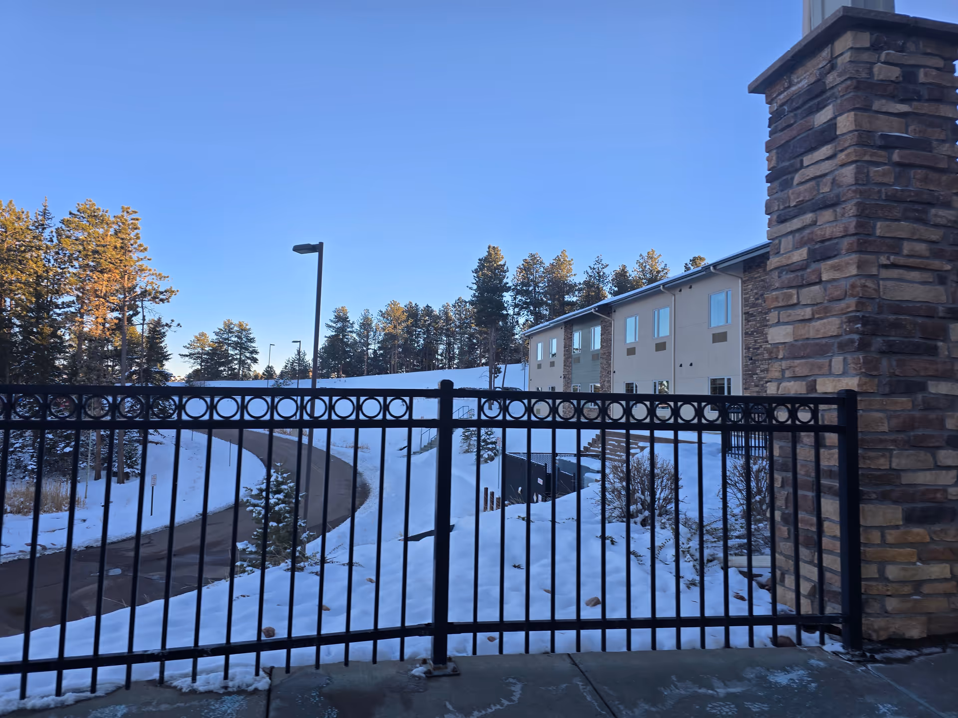 Snow-covered driveway and black iron fence in front of a two-story building with pine trees and a clear blue sky.